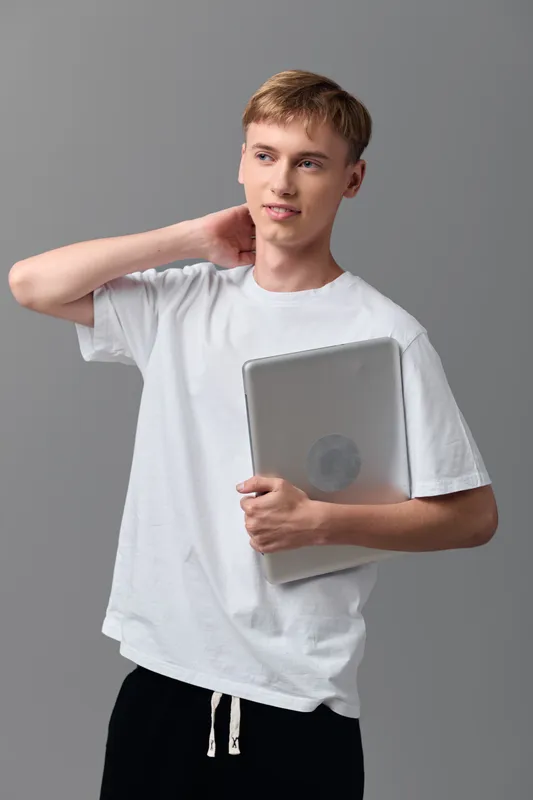 Casual young male holding tshirt and laptop in indoor studio with white background, showcasing modern learning and technology setup for education, work and everyday tasks.