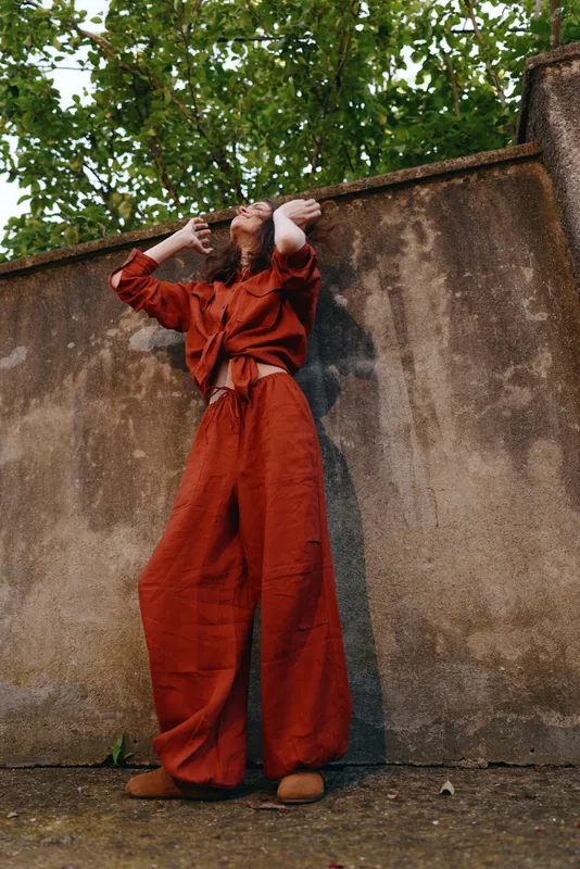 A woman in a red orange jumpsuit leans against a weathered wall outdoors, hands shielding her eyes as sunlight filters through trees. Fashionable street style portrait capturing resilience