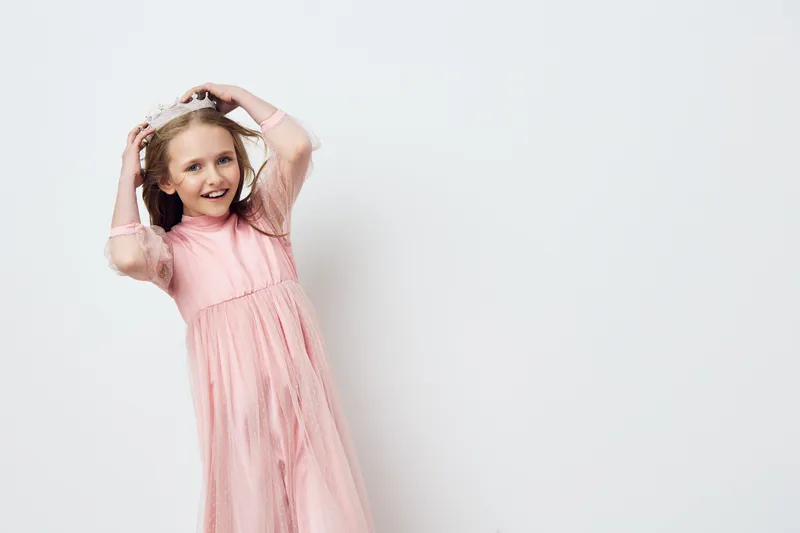 Cheerful young girl in a pink dress, playfully posing against a light gray background, radiating joy and innocence, perfect for children s fashion and lifestyle imagery