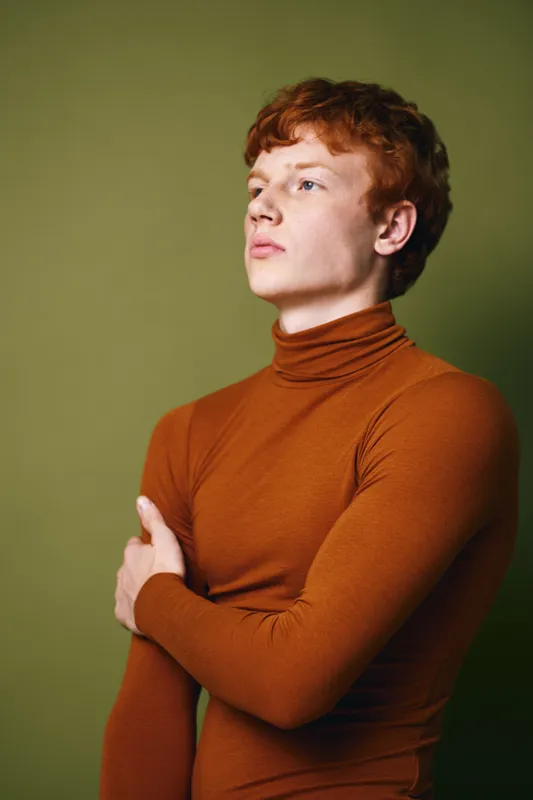 Young man with red hair and fair skin posing confidently in brown turtleneck sweater against green background. Portrait, fashion, casual style and thoughtful expression.