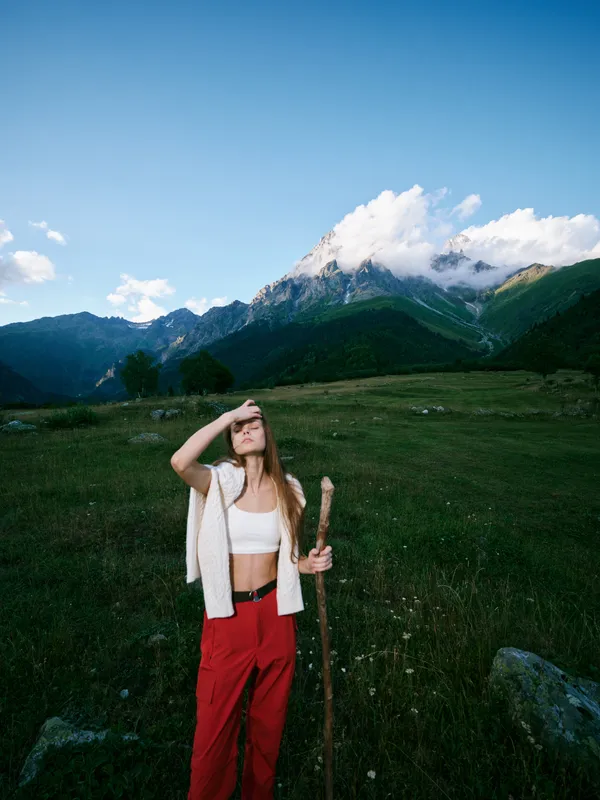 outdoor hiking mountains landscape woman staff meadow sky A woman stands in a grassy field with a walking staff, wearing a white top and red pants, against dramatic peaks, conveying exploration and