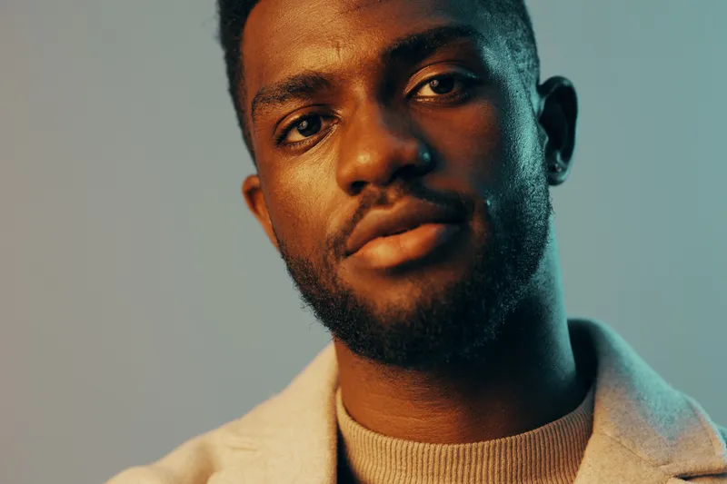 Confident young African man in stylish attire with soft lighting, showcasing a cool expression against a minimalist blue backdrop