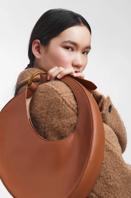 asian woman with a round brown leather bag and cozy wool coat stands in a studio, exuding confidence and style while showcasing a fashionable accessory moment.