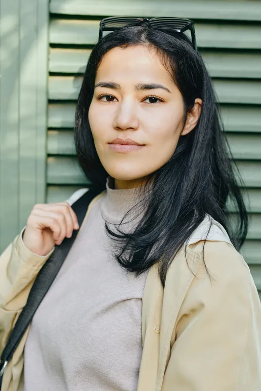 Woman in casual outdoor fashion wearing a beige coat and sweater, sunglasses on head, standing beside green shutters, conveying confident streetstyle and everyday chic in natural light, urban vibe