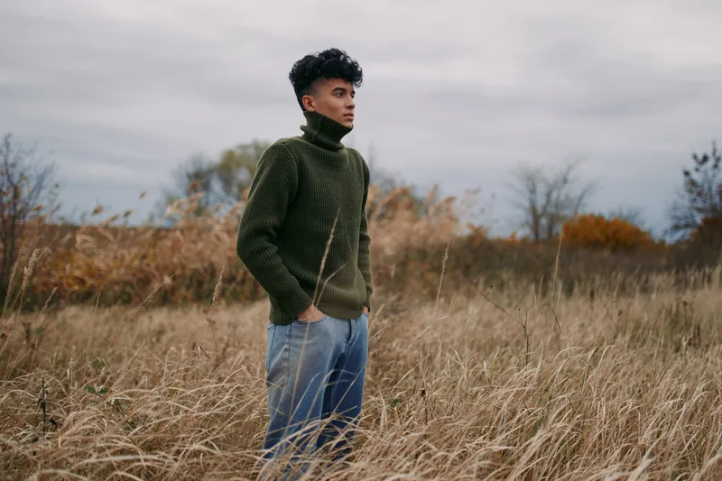 A young man stands in tall dried grass wearing a chunky knit turtleneck, gazing thoughtfully; image highlights natural pose, authenticity and reliability in candid outdoor portrait.