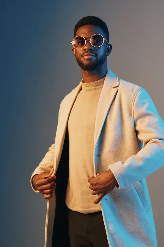 Stylish young man wearing a beige coat and sunglasses, showcasing confidence against a gradient blue background, perfect for fashion and lifestyle themes