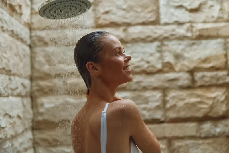smiling woman enjoying outdoor shower with stone wall background, wet hair, natural light, and relaxed summertime atmosphere, health and wellness concept