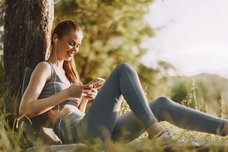 Young happy woman enjoying music on her smartphone outdoors, wearing activewear, surrounded by nature in a sunny setting, showcasing a relaxed lifestyle