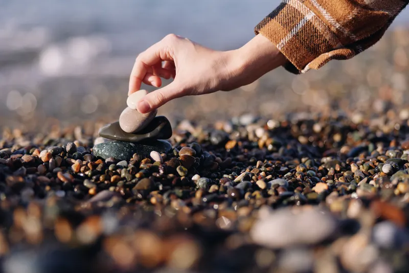 Closeup of a hand placing a pebble on a beach of small stones, with blurred water in the background, emphasizing texture, balance and mindful outdoor action.