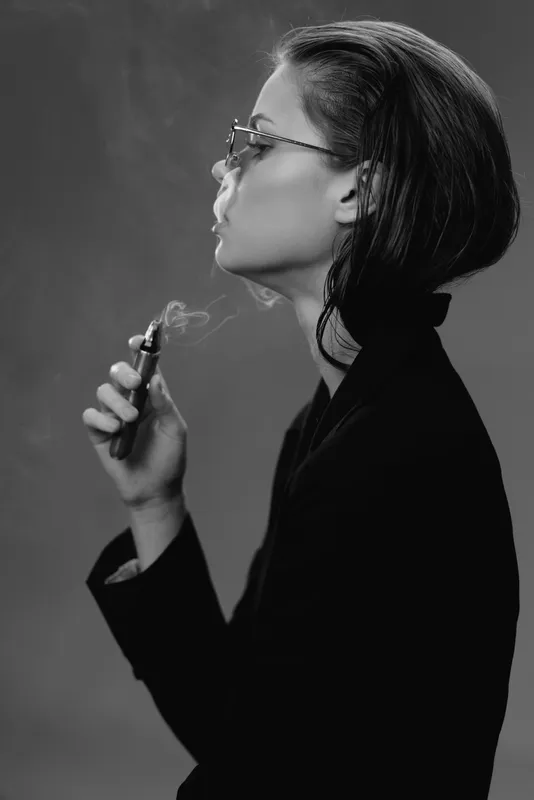 Black and white portrait of a thoughtful woman smoking with glasses in a dark outfit.