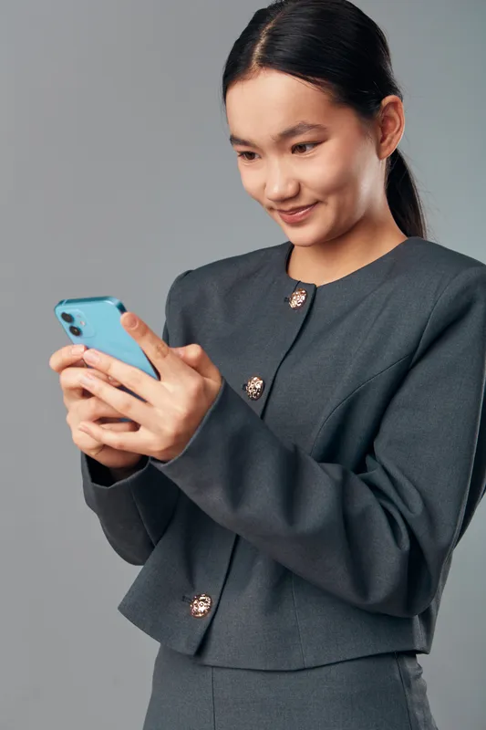 Woman with smartphone texting message and smiling, business outfit with dark jacket alone on grey background. Asian female holding phone, communication and technology concept for smartphone usage.