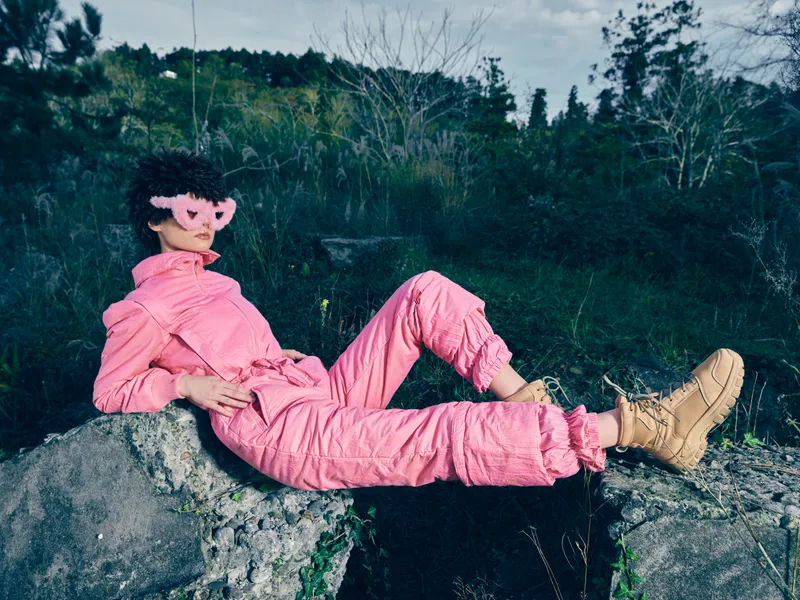 Pink jumpsuit, masked figure outdoor on rock, relaxing pose amid nature. Surreal fashion scene with bold color against rugged scenery, capturing quiet solitude and playful mystery in an outdoor