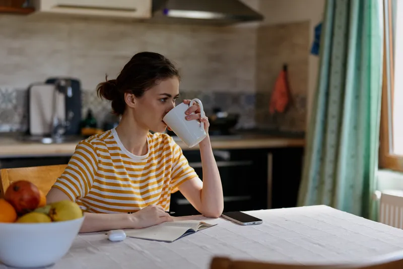 Woman at home sits at a kitchen table with a mug, notes and tablet, wearing a striped shirt, warm morning light, casual lifestyle, calm focused moment in an everyday setting