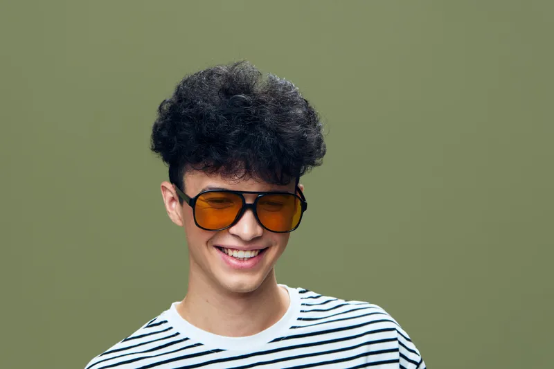 smiling man wearing retro orange sunglasses and striped shirt against plain olive background, casual style, youthful and confident expression