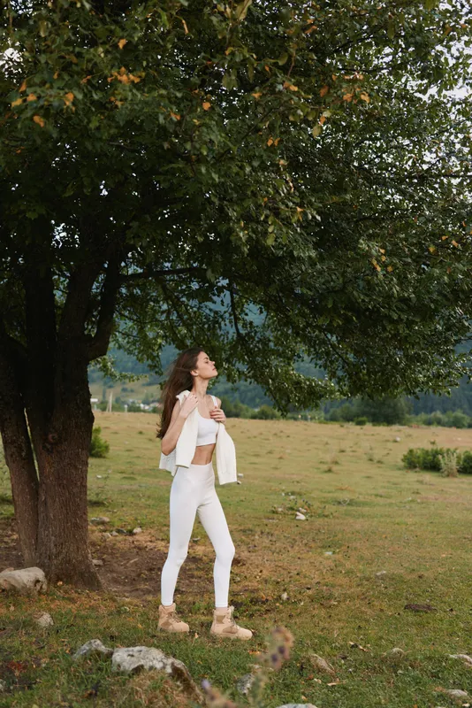A stylish woman in white outfit stands under a large tree in an open park, enjoying a sunny day. Casual mood, feminine fashion, and outdoor living.