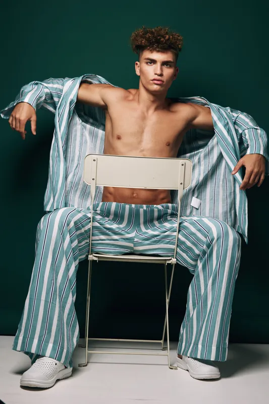 Young man sits on white metal chair shirtless wearing wide striped blue and white pants and open oversized long sleeved shirt in studio against dark green background with confident relaxed posture.