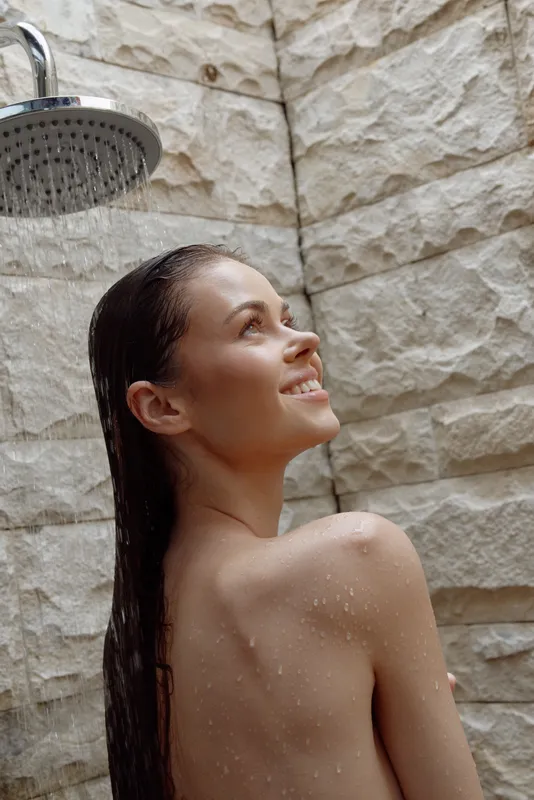 smiling woman enjoying outdoor shower with wet hair against textured stone wall, natural light enhancing fresh and relaxed mood in summer setting