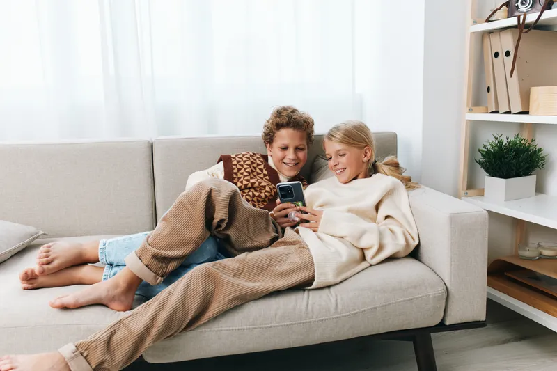 Two children lounge on a light sofa, smiling as they share a smartphone in a bright living room. Soft lighting, cozy attire, and a candid, relaxed indoor moment.