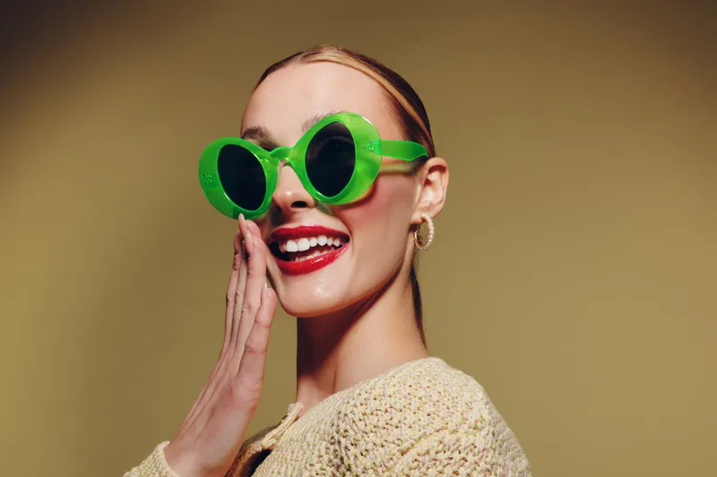 Smiling woman with bright red lipstick wears large green sunglasses and beige sweater, posing with hand near face on yellow background. Concept of fashion, style, and joyful expression.