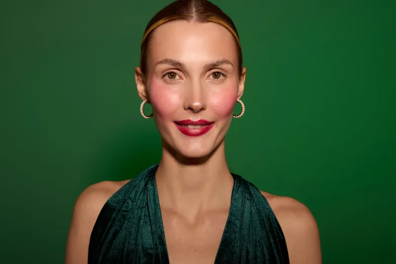 Portrait of confident woman with red lipstick and hoop earrings wearing dark green dress against green background. Studio lighting highlights natural skin and elegant style.