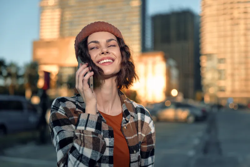 Woman on phone smile in urban street at golden hour glow, candid lifestyle portrait conveying authenticity, joyful expression and mindful living with emotional storytelling mood.