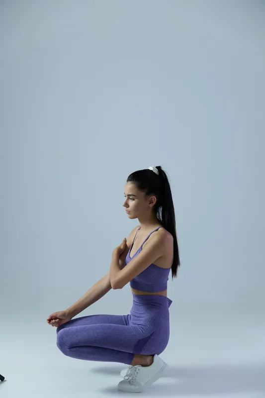 Young woman sport fitness dressed in purple activewear squatting on a light background, focused and calm, indoor workout concept, health and wellness.