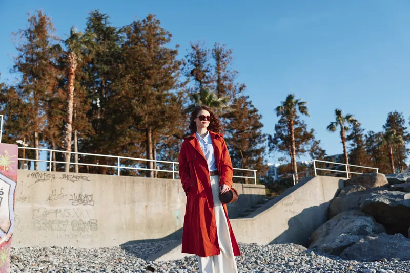 Smiling woman in a red coat standing on a rocky beach with trees in the background. Fashionable outerwear, sunny outdoor scene, confident pose and relaxed mood.