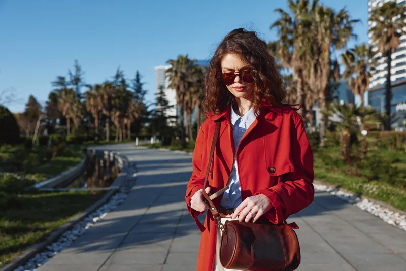 Stylish woman with curly hair wearing a red coat walks along a sunny promenade with palm trees, sunglasses on, carrying a brown bag, exuding confidence.