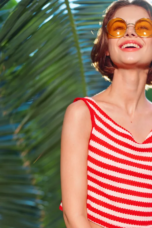 A cheerful woman wearing a red and white striped top stands outdoors with palm leaves in the background, wearing round sunglasses and smiling broadly, radiating sunny summer energy.