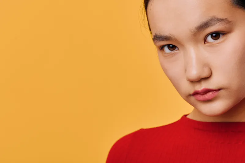 Portrait of young woman with serious expression wearing red top against yellow background. Closeup of female face with natural makeup and direct eye contact for emotion or mood.