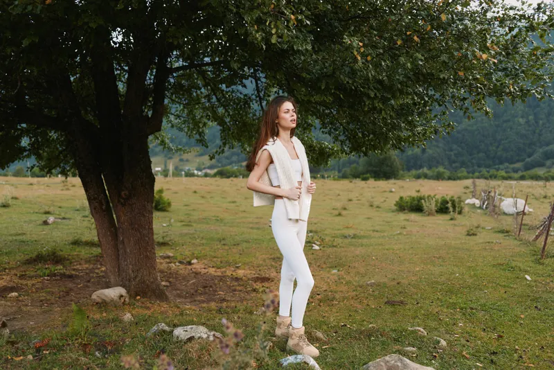 Young woman in white outfit standing outdoors near tree in grassy field, relaxed posture, natural light, casual lifestyle, peaceful rural environment, fresh air