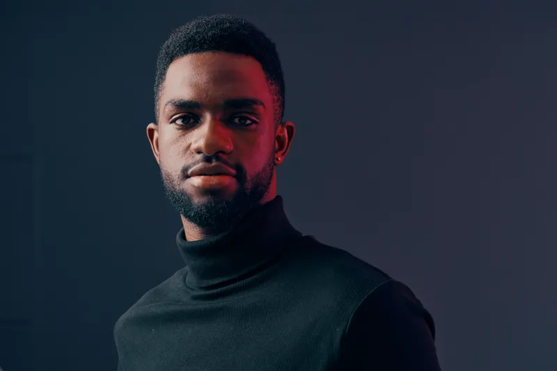 Confident young African man wearing a black turtleneck sweater, showcasing a serious expression against a dark background with contrasting red highlights, ideal for fashion and lifestyle concepts