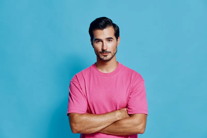 Confident young man in pink t-shirt standing with arms crossed against vibrant blue background, looking at camera with a serious expression. People lifestyle concept.