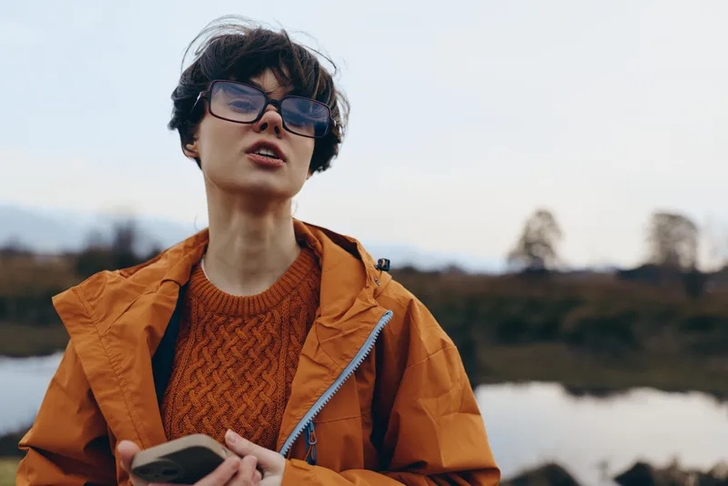 Woman in outdoor lifestyle wearing orange jacket and glasses, holding notebook while looking ahead, breathing fresh air near river and hills, soft overcast light, calm travel mood today