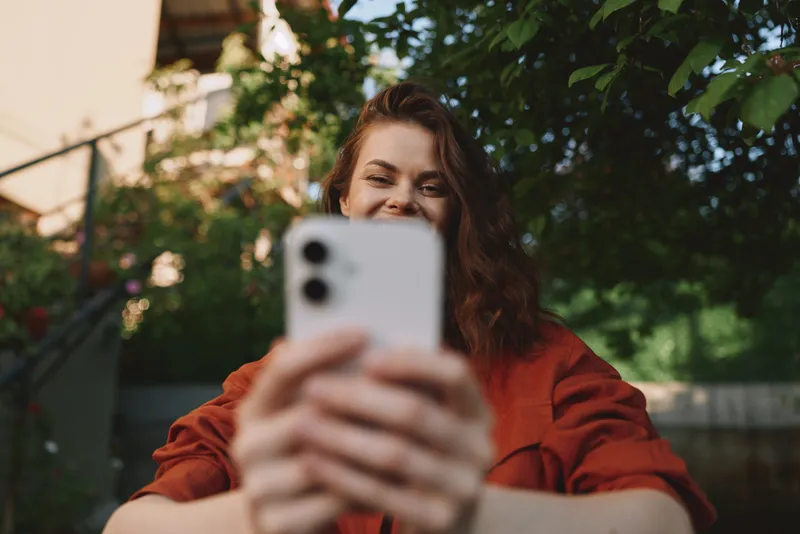 Woman lifestyle outdoors, taking a selfie with a phone, smiling at the screen, casual pose in a sunny garden, natural light and relaxed mood, for social media sharing