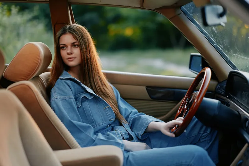 Young woman in denim clothing sitting in a vintage car, conveying a relaxed and stylish vibe in a natural outdoor setting.