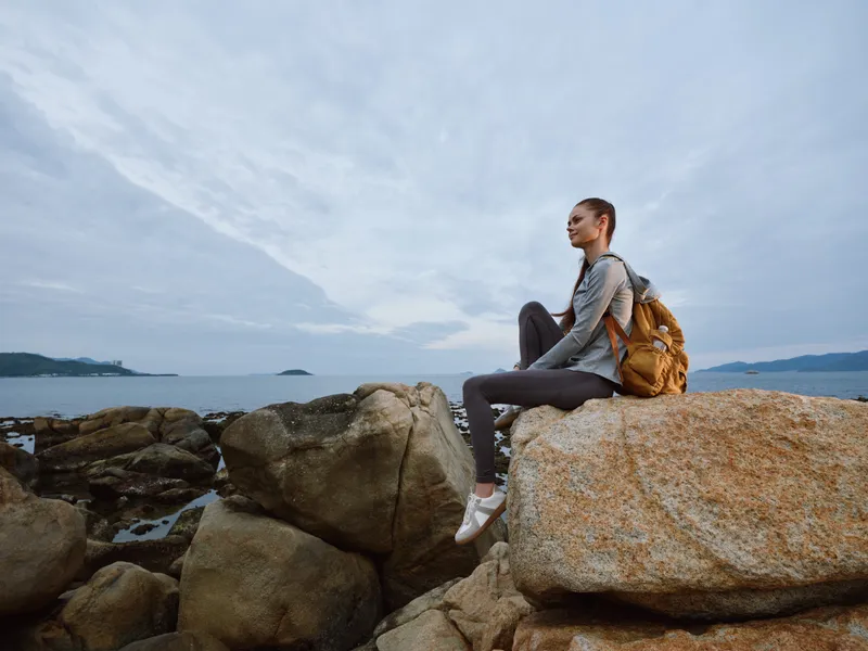 Young woman in outdoor sportswear sitting on rocks by the sea, showcasing an adventurous spirit and connection with nature, perfect for travel and fitness themes