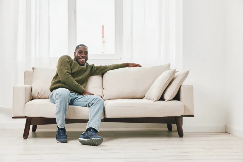 Happy Black man in cozy sweater on beige couch, showcasing comfort and relaxation in a bright, modern living room environment