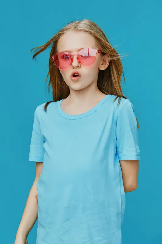 A young girl with long hair wearing a blue t shirt and stylish pink sunglasses, expressing joy against a vibrant blue background