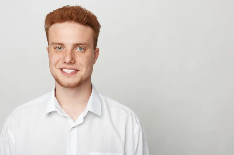 Smiling young man with red hair and freckles wears white shirt, looking at camera on light background. Concept of casual style, confidence, youth, and natural portrait photography.