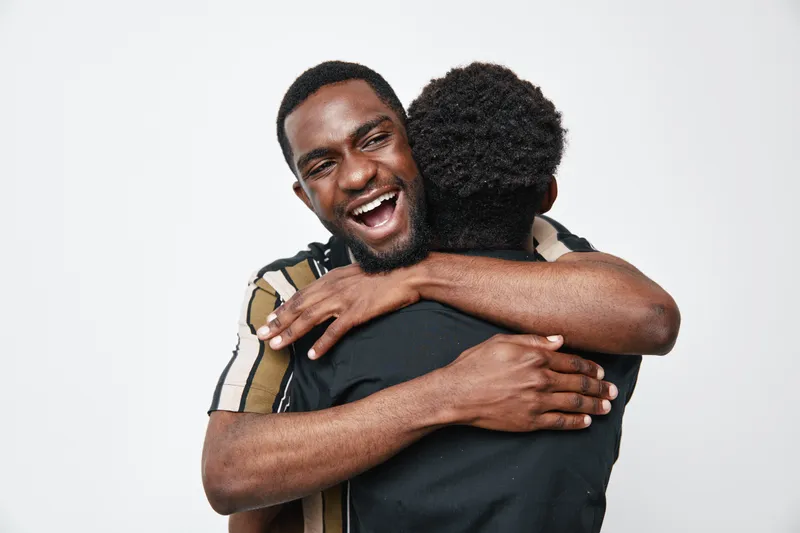 Two black men with dark skin hugging each other on white background. African ethnicity, friendship and happiness with smiling face and casual clothing in studio.