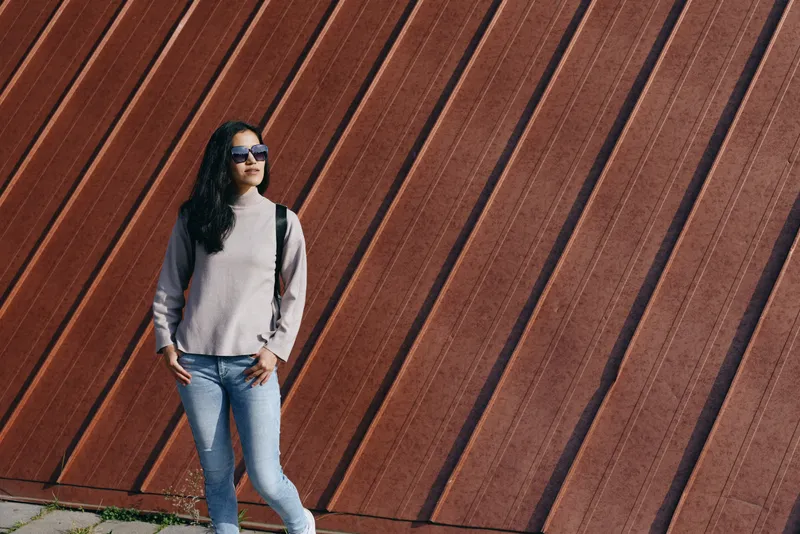 Woman wearing sunglasses, sweater and denim jeans stands against a diagonal wooden wall outdoors, casual street style, contemporary fashion, urban backdrop and confident gaze