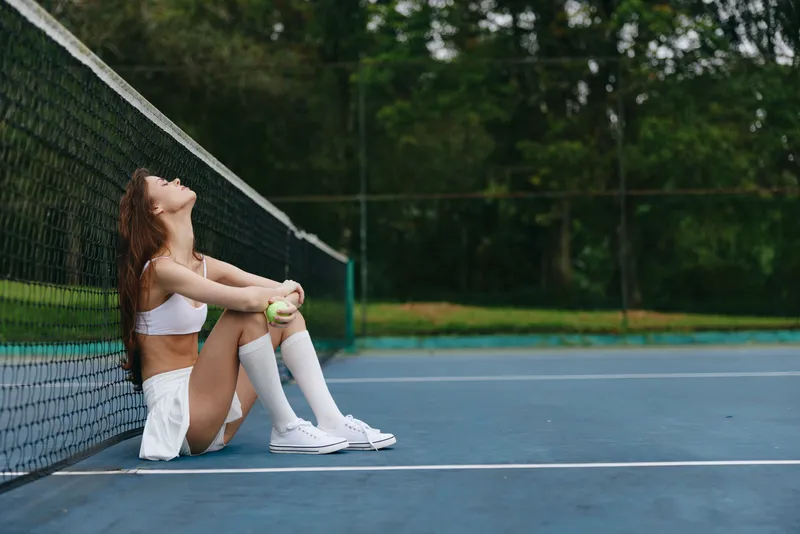 Young female tennis player in sportswear, expressing relaxation and determination, sitting on the court, surrounded by vibrant greenery