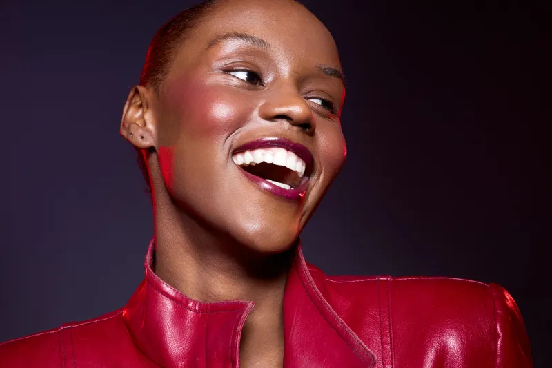 Smiling African American woman with dark skin wearing a red leather jacket laughs joyfully against a dark purple background. Concept of happiness, fashion, and beauty with vibrant colors.