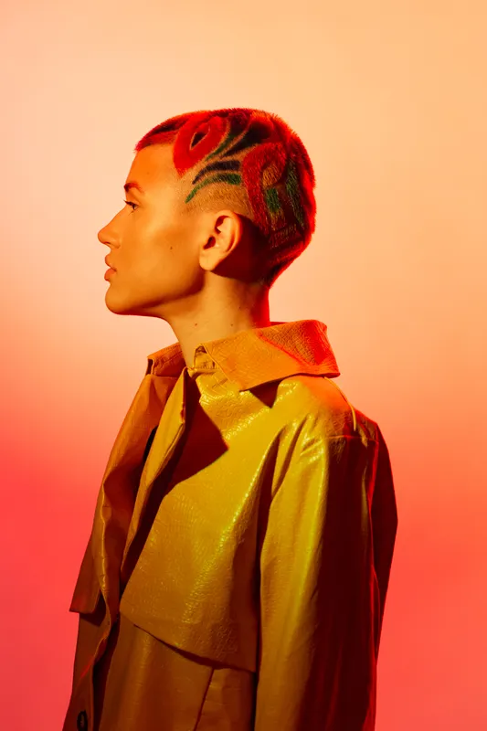 Woman with artistic short hair dyed red and green, wearing a yellow coat, standing in profile against a warm orange background, showcasing modern fashion and creativity.