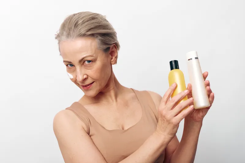 Mature woman with short hair holds two cosmetic bottles, one yellow and one white, against a plain white background. Concept for skincare, beauty, wellness, and personal care routines.