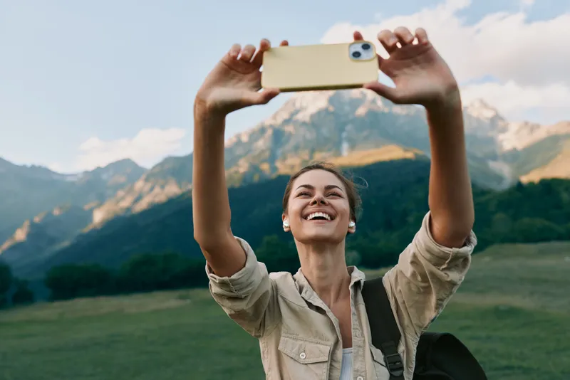 Young woman in a light jacket takes a cheerful selfie in a green field with distant mountains, capturing a carefree outdoor moment