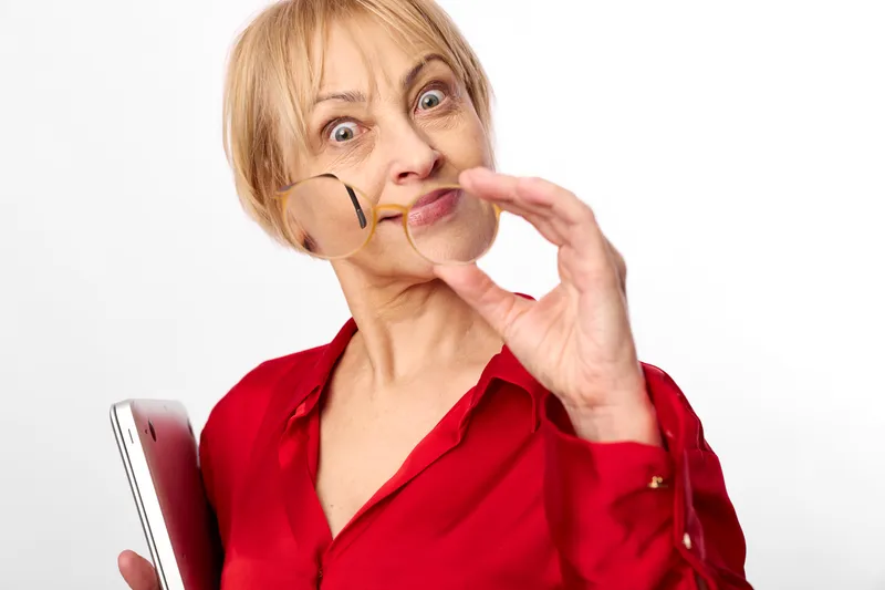 Senior woman in red shirt holding smartphone, adjusting glasses with finger, surprised playful face, looking at camera, white background close up portrait