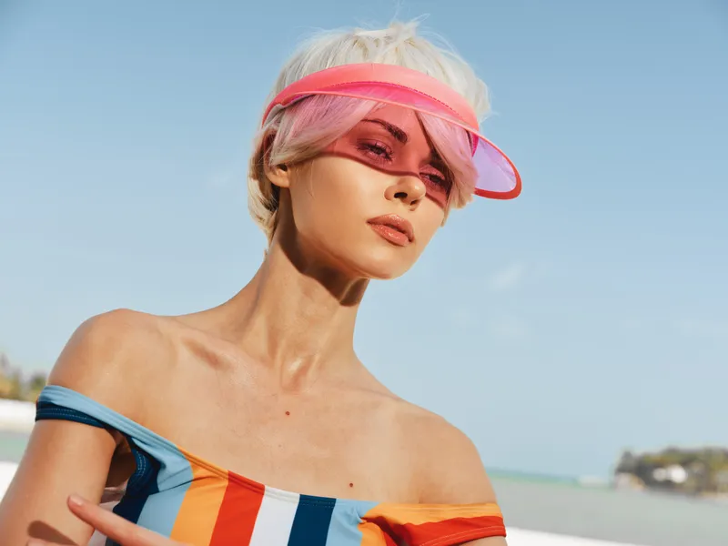 Closeup portrait of a stylish model on a sunny beach, wearing a pink visor and a colorful offshoulder top, dramatic lighting and a confident expression under clear blue sky.