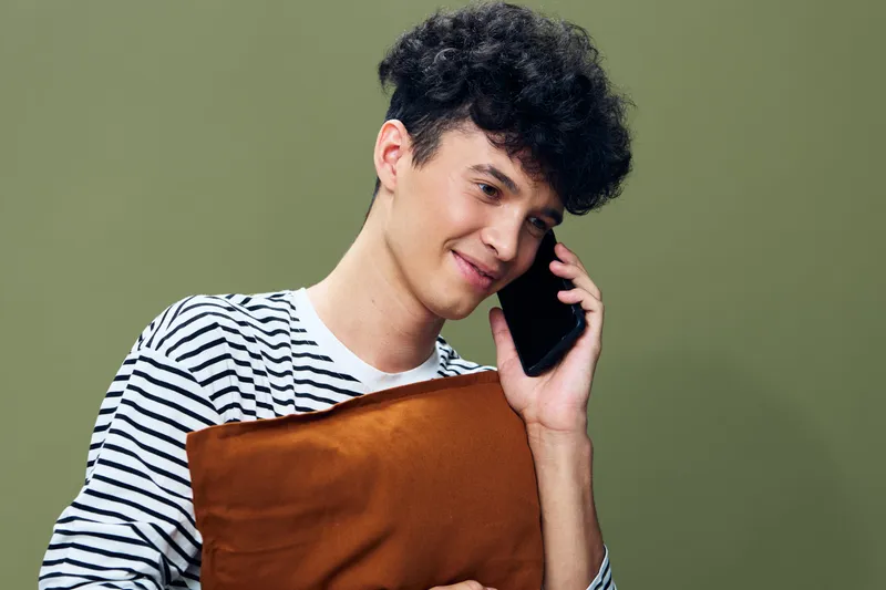 young man talking on smartphone holding brown pillow, casual striped shirt, smiling with curly hair against green background, relaxed and cheerful mood
